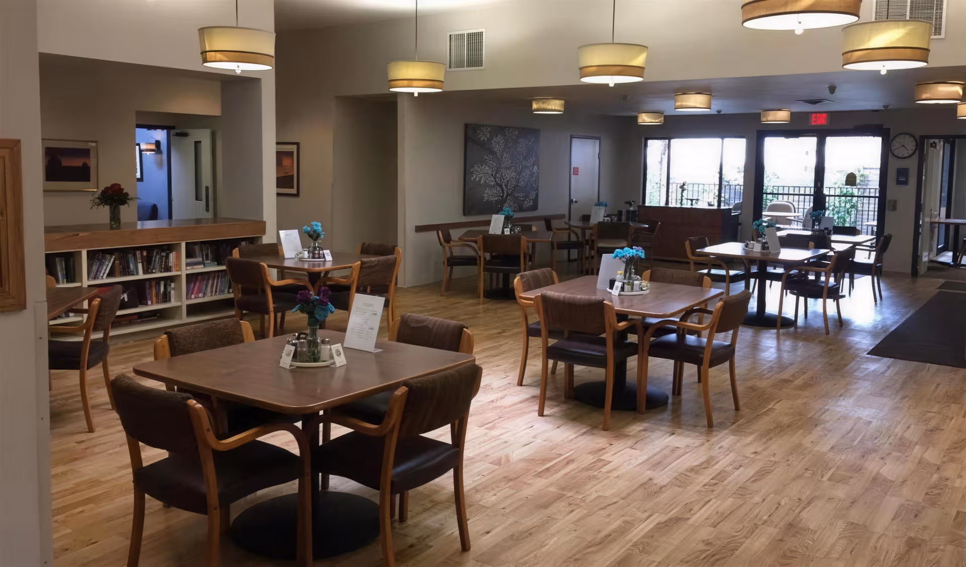 Bright senior living dining area with wooden tables, chairs, bookshelves, and pendant lighting.