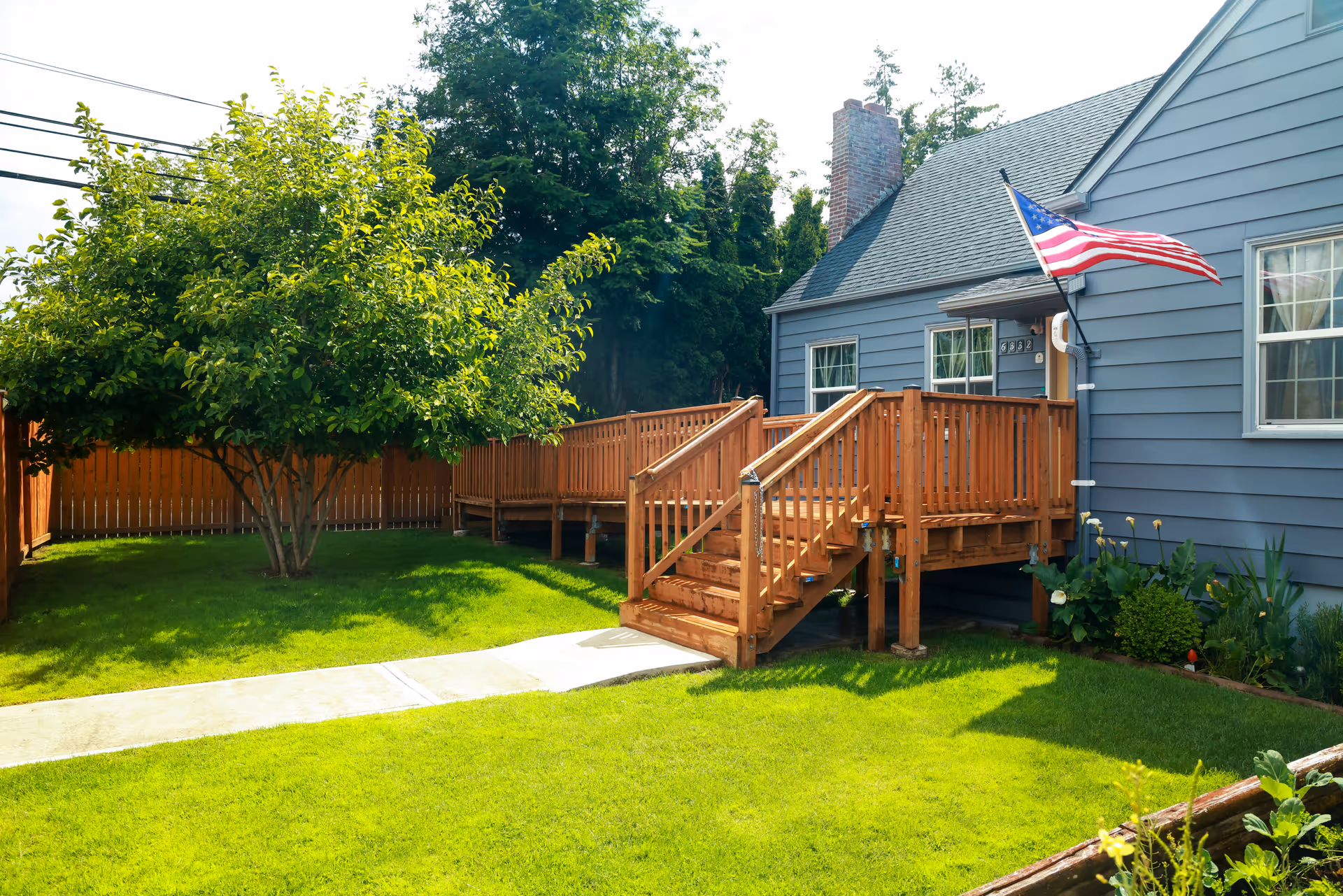 Front yard of a blue house with a wooden deck and American flag.
