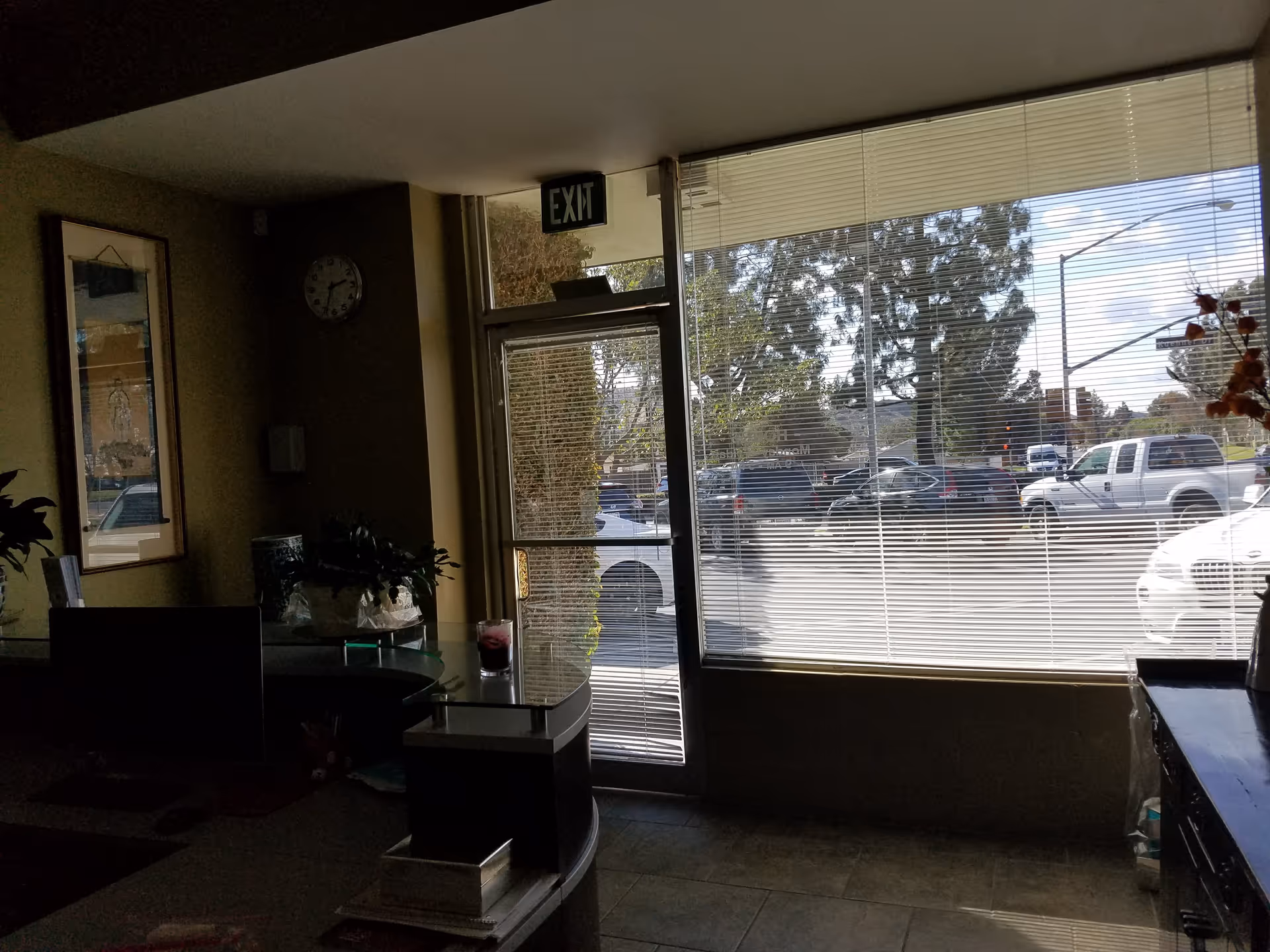 Interior lobby with glass entrance, blinds, plants, exit sign, and street view outside.