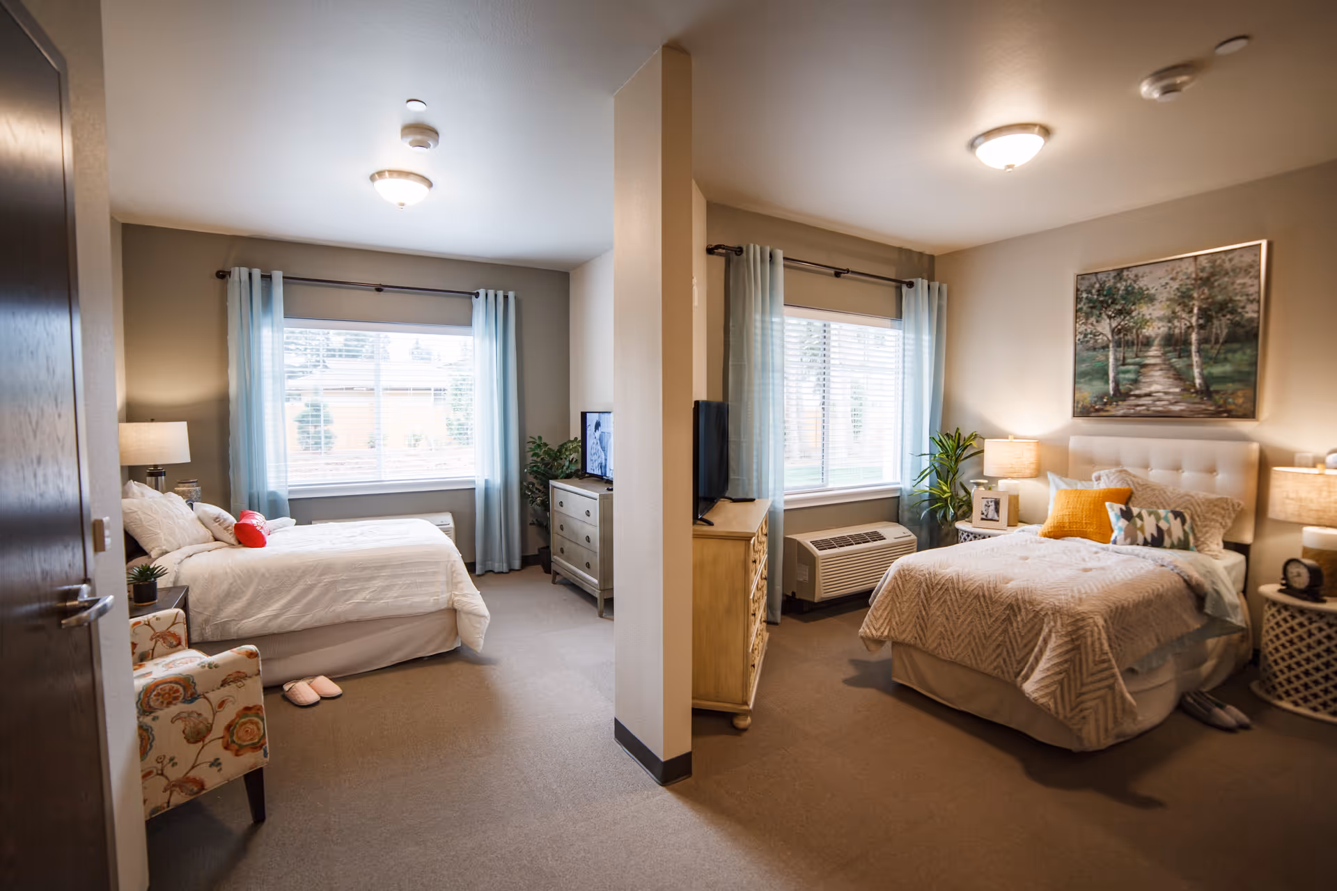 A cozy senior living bedroom area divided by a partial wall, featuring two beds with white bedding, bedside tables with lamps, a floral armchair, a dresser with a TV, large windows with light blue curtains, and decorative plants and artwork.