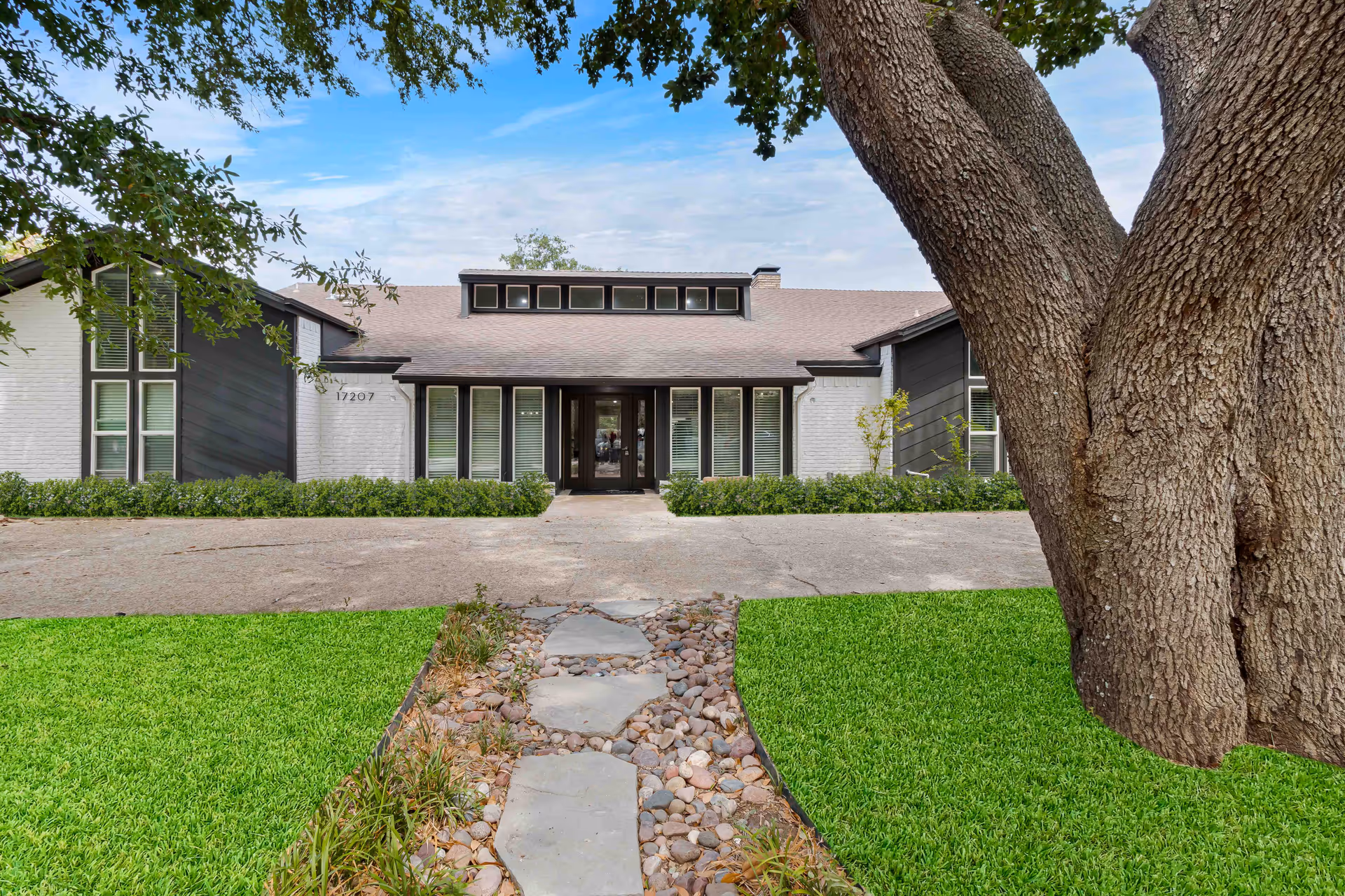 Front exterior view of a single-story building with white brick and dark wood siding, large windows, and a central entrance with glass doors. A stone pathway leads through a green lawn and landscaping to the entrance. A large tree is visible on the right side of the image.