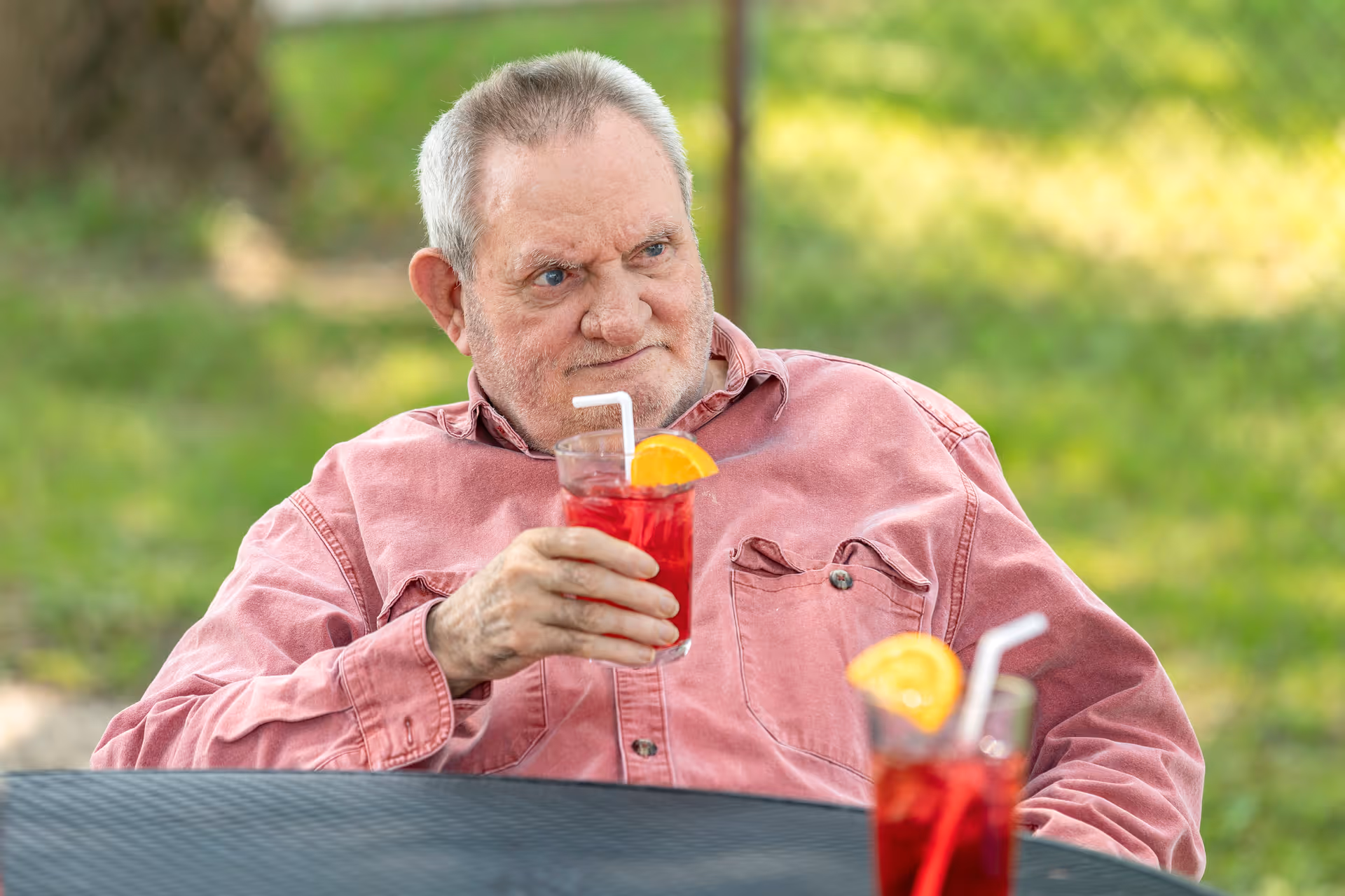 An elderly man wearing a pink button-up shirt is sitting outdoors at a black metal table, holding a glass of red beverage garnished with an orange slice and a white straw. Another similar drink is on the table in front of him, with a blurred green background.