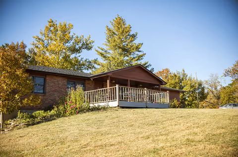 Brick senior living building with a wraparound wooden deck on a grassy, tree-lined hill.