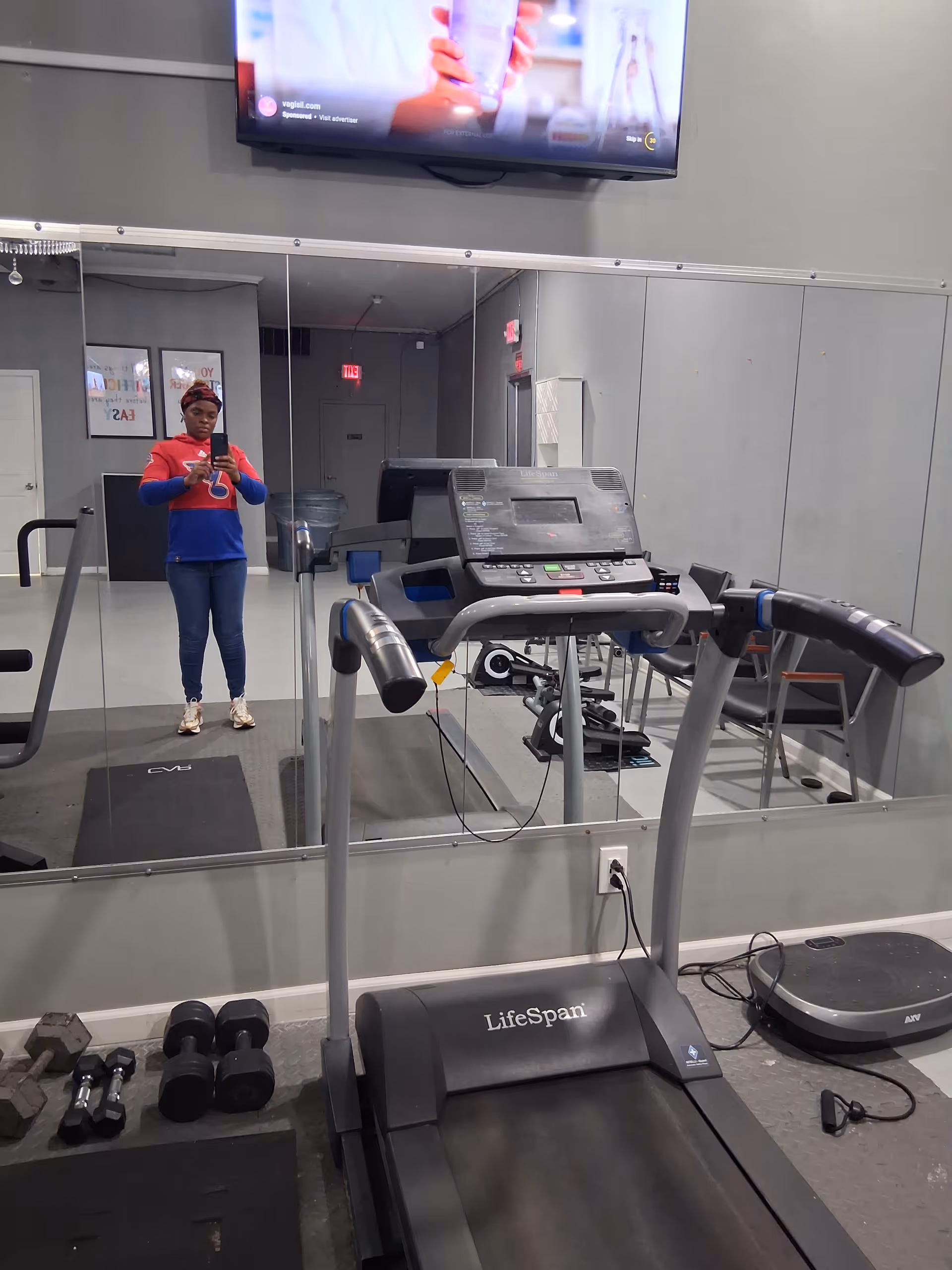 Person stands in a gym area with treadmill, dumbbells, and mirrored wall.