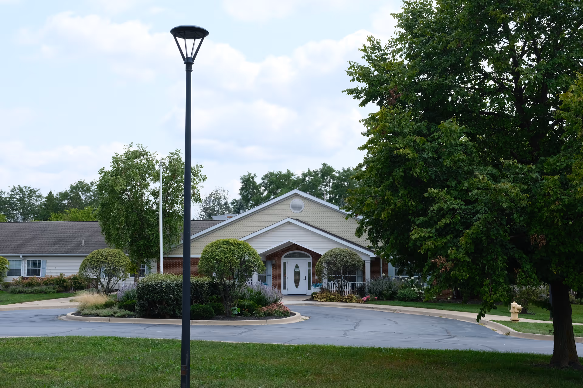 Front entrance of a single-story senior living building with a circular driveway, landscaping, and a lamp post.