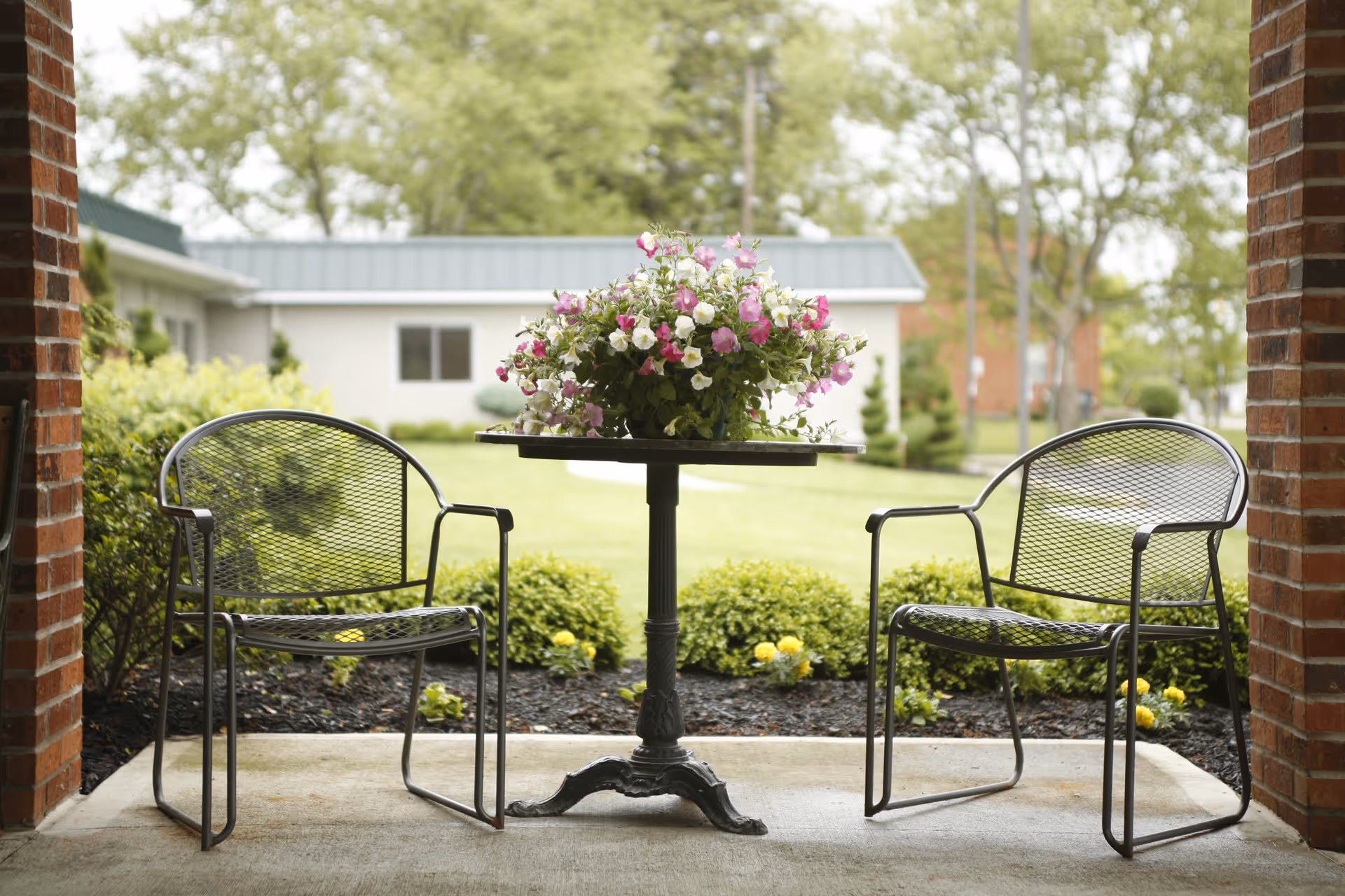 Two metal chairs flank a small round table with flowers on a covered outdoor patio.