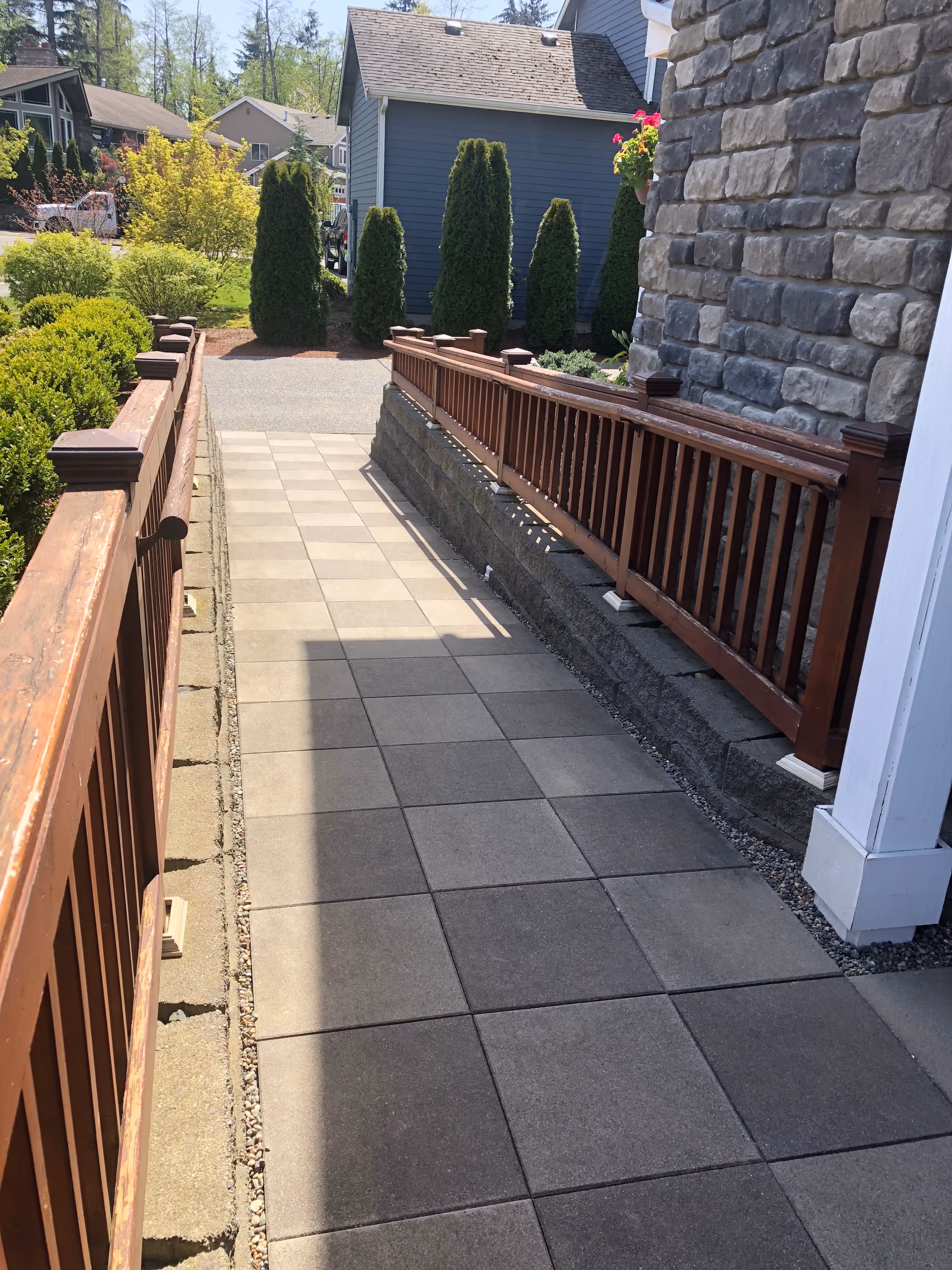 Sunlit outdoor walkway with brown railing, stone wall, and manicured shrubs outside a senior home.
