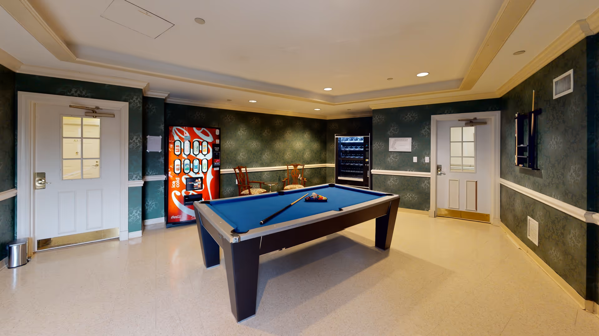 A recreational room with a blue felt pool table in the center, a pool cue resting on it, and a rack of billiard balls arranged for a game. The room has green patterned wallpaper with white trim, two white doors with windows, two wooden chairs with cushions, a red Coca-Cola vending machine, and a black vending machine. A wall-mounted pool cue rack is visible on the right wall.