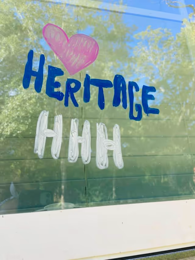 Window sign with blue HERITAGE and a pink heart above white HHH letters on glass.