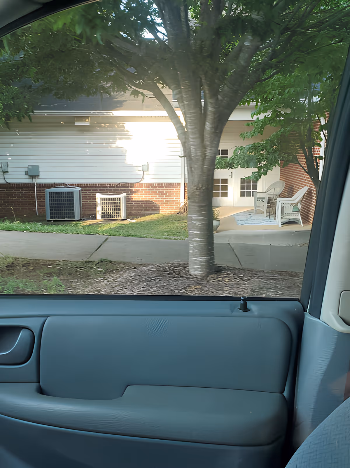 View from inside a parked car showing a tree, sidewalk and the exterior patio of a senior living building with chairs.