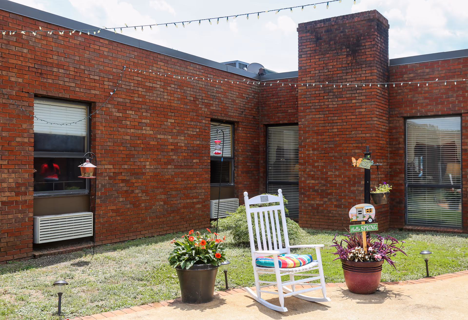 Brick courtyard of a senior living facility with a white rocking chair, planters, and string lights.