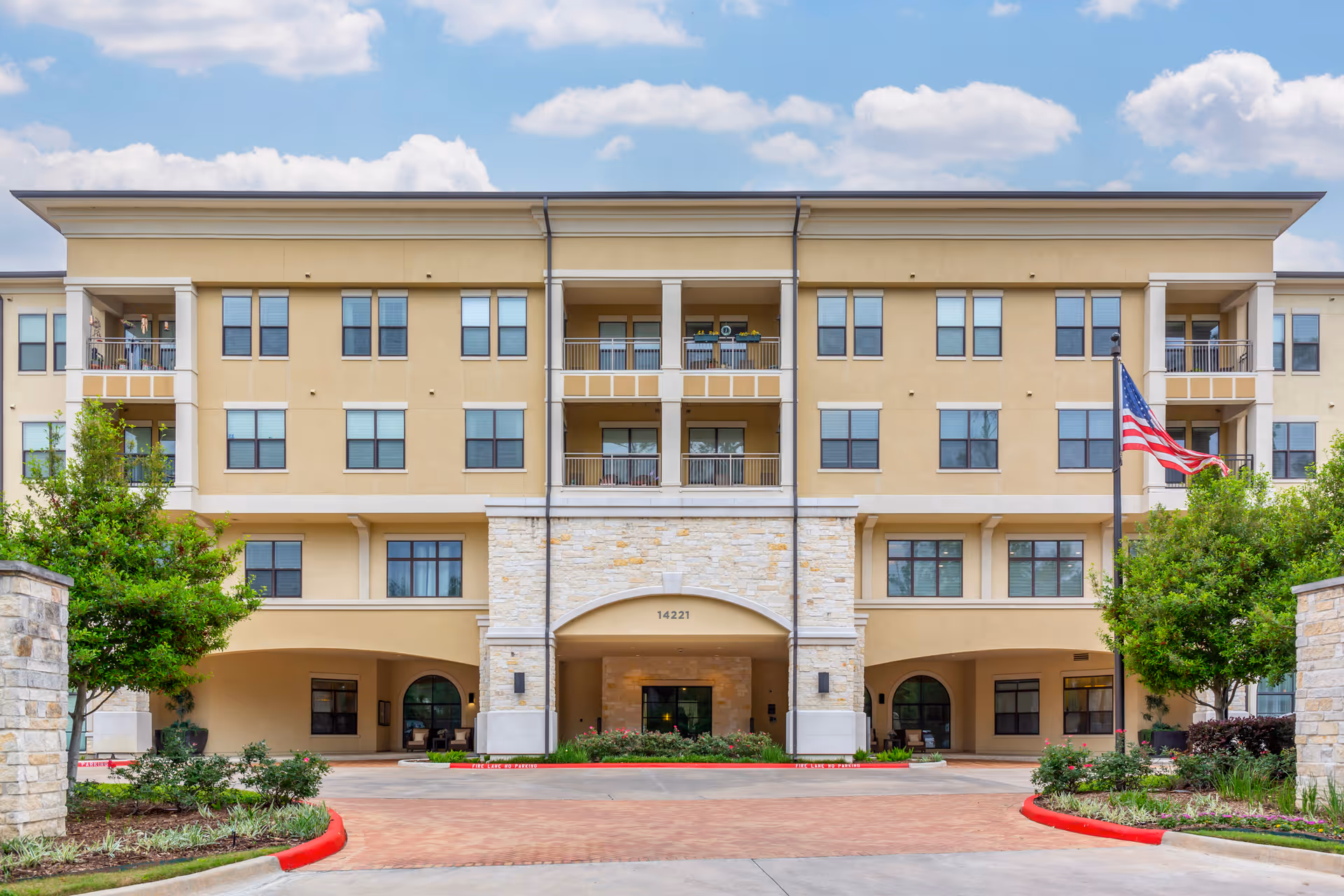 Front exterior view of a multi-story senior living facility building with beige walls, multiple windows, balconies, an American flag on a flagpole, and landscaped greenery in front under a partly cloudy sky.
