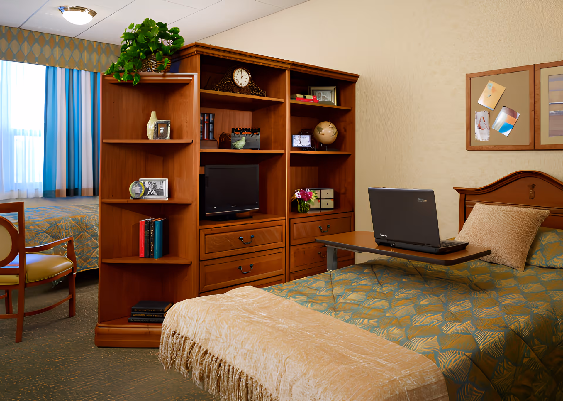 A cozy bedroom in Alden Estates of Naperville featuring a bed with a green and gold patterned bedspread, a beige throw blanket, and a pillow. A wooden headboard is visible along with a small overbed table holding a laptop. Behind the bed is a wooden shelving unit with various decorative items including a clock, globe, books, and framed photos. A window with blue and white curtains and a wooden chair are also visible.