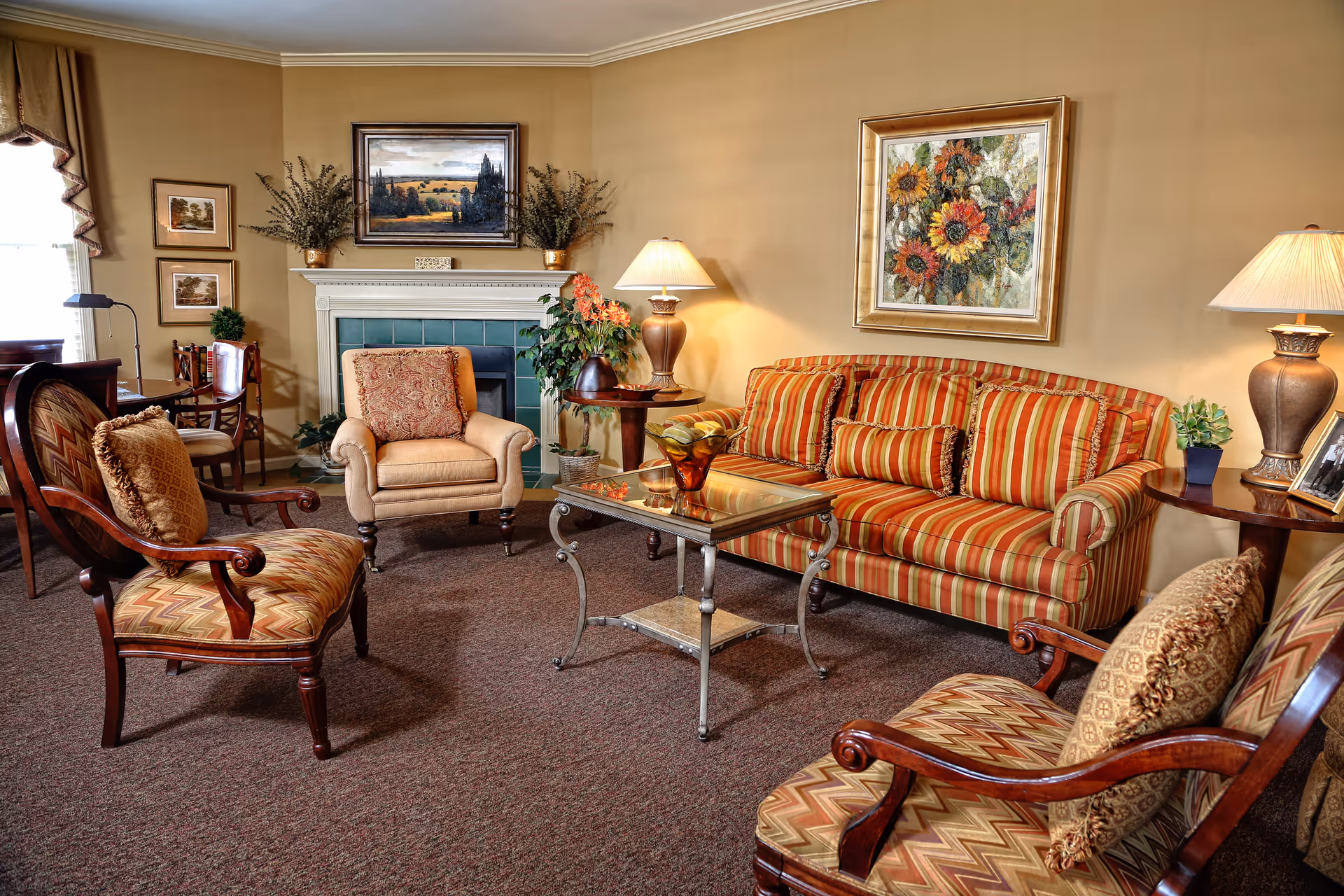 A cozy living room with a striped sofa, two patterned armchairs, and a beige armchair arranged around a glass coffee table. The room features a fireplace with green tiles, framed paintings on the walls, table lamps, and decorative plants.