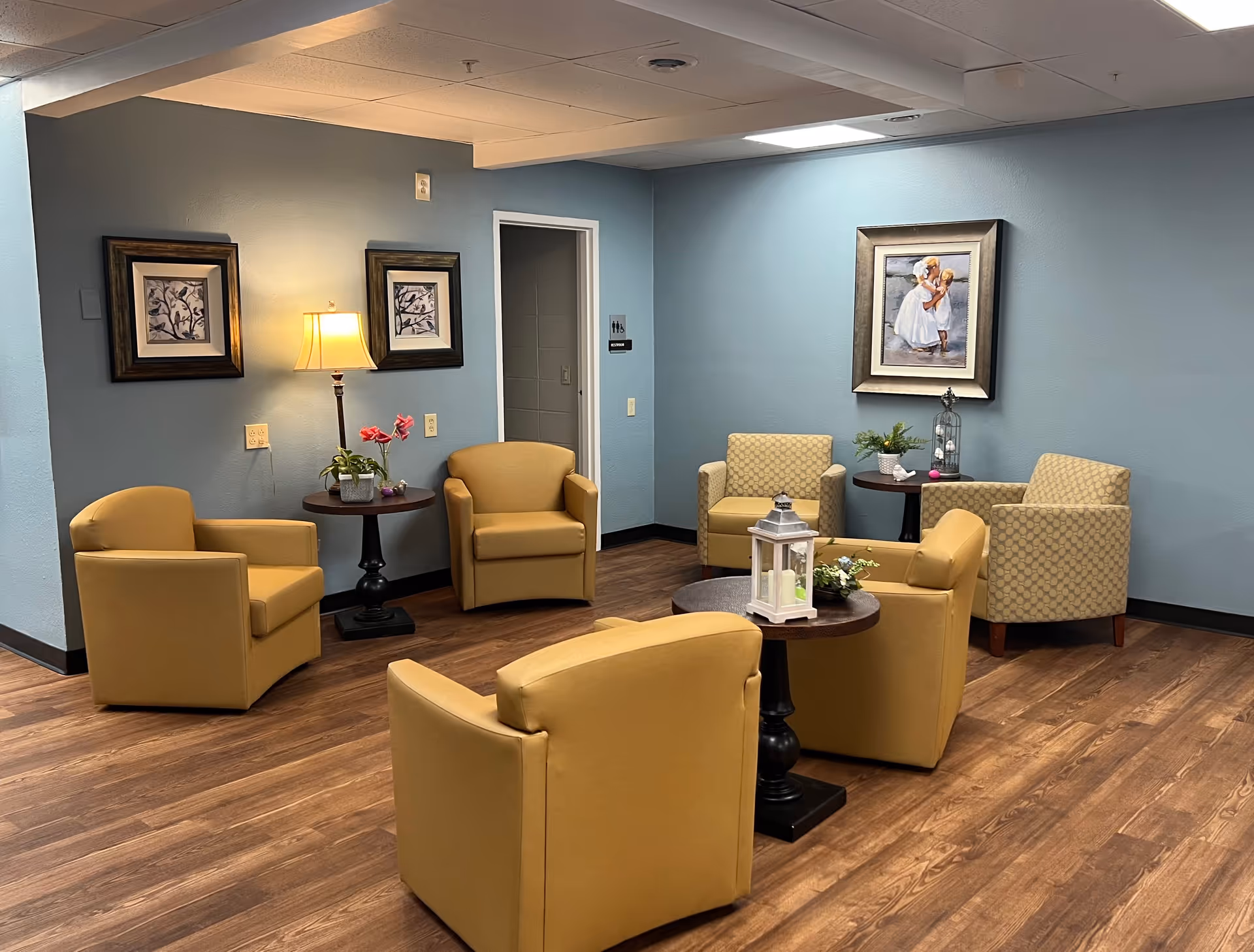 A spacious lounge with mustard chairs, small tables, and framed art on blue walls.