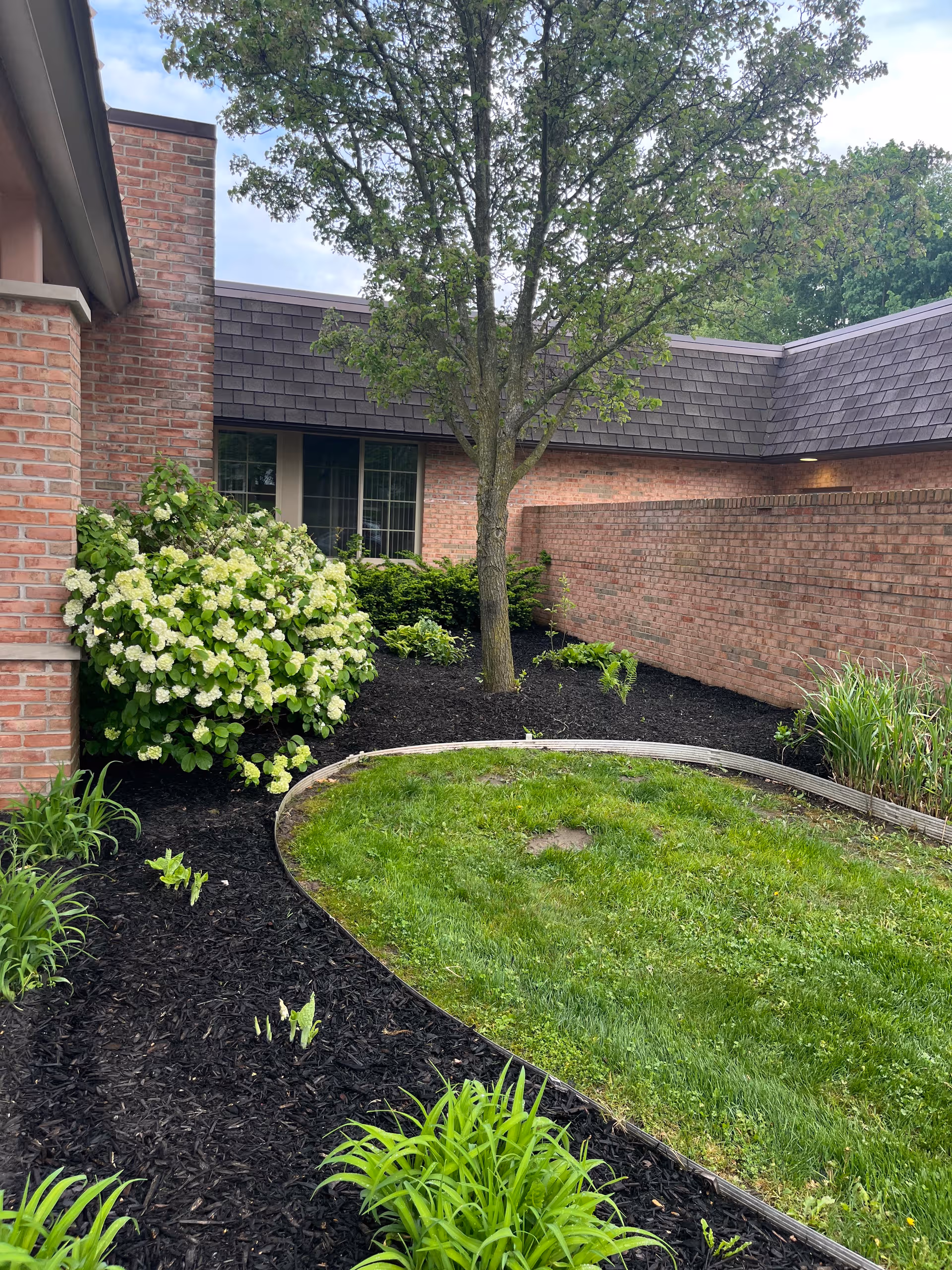 Brick nursing facility exterior with a landscaped courtyard, tree, and curved mulched beds.