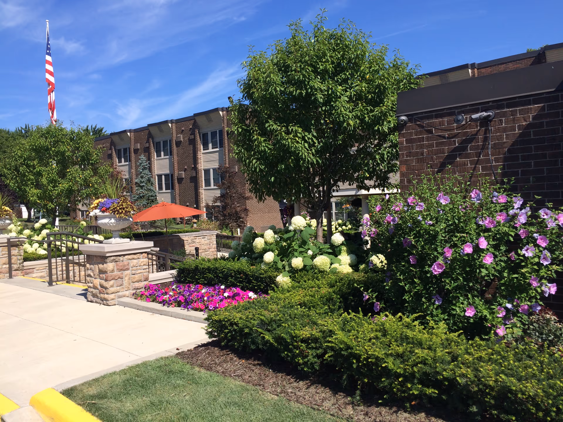 Front entrance of a brick senior living building with landscaped flowerbeds, trees, and an American flag.