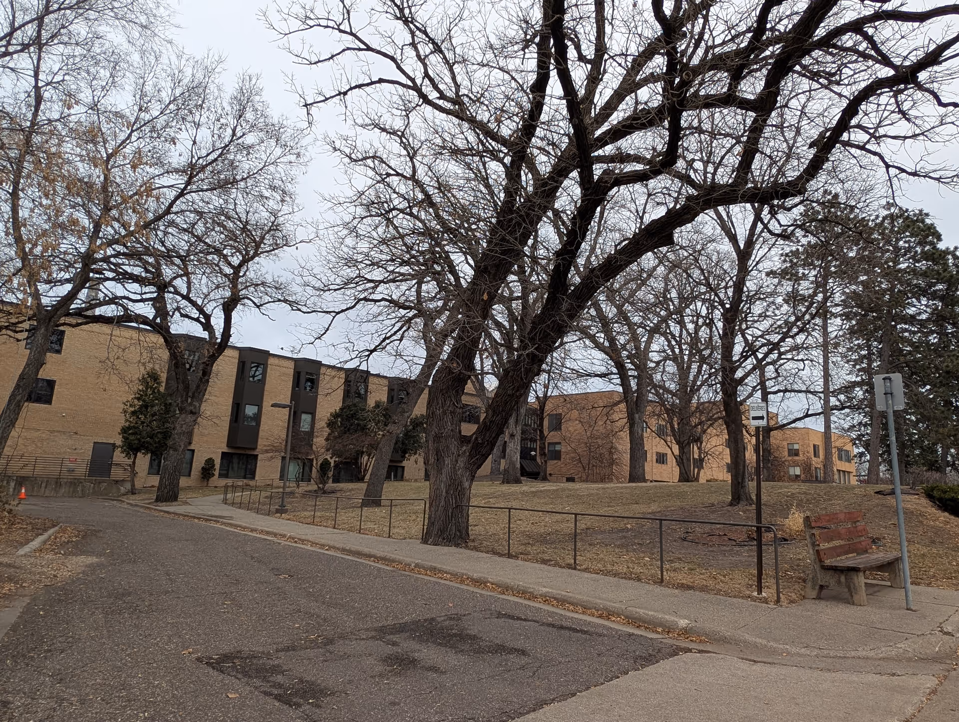 Brick multi-story senior living building set behind leafless trees with a sidewalk and a bench in the foreground.