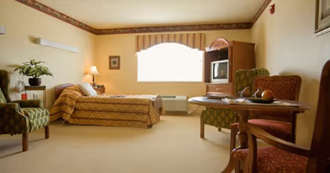 A furnished senior living suite with a bed, sofa, table, and television in soft lighting.
