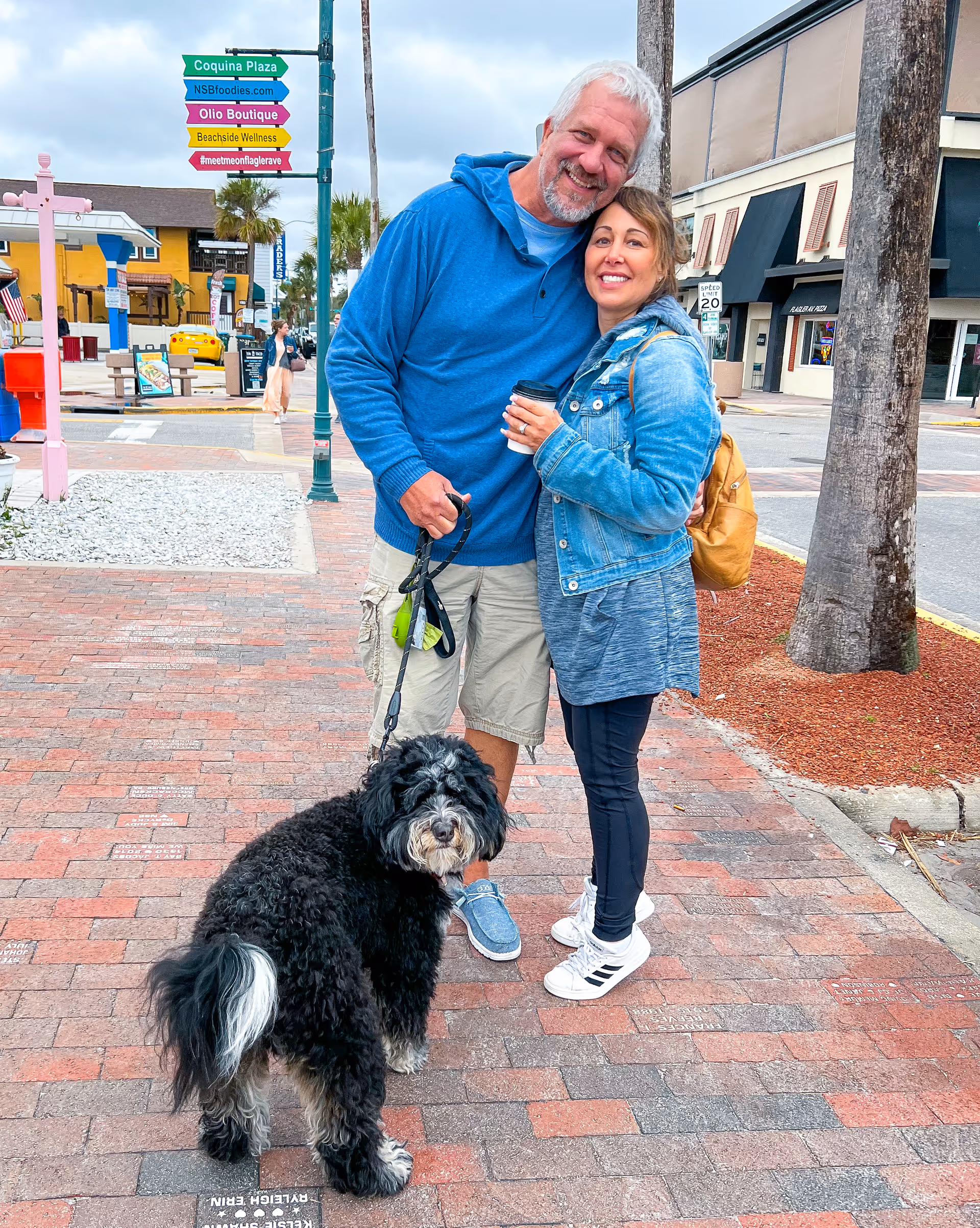 Smiling couple with a leashed black dog on a brick sidewalk in an outdoor shopping area