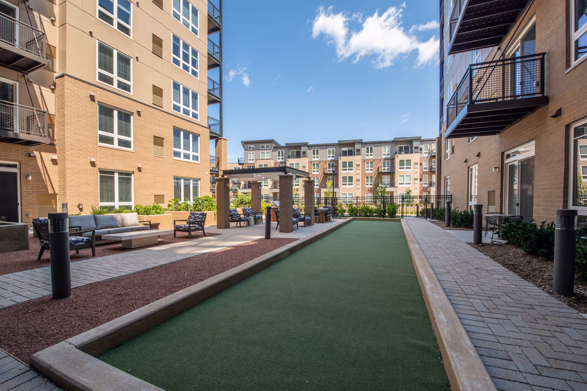 Sunny outdoor courtyard between apartment buildings featuring a bocce court, seating areas, pergola, and balconies.