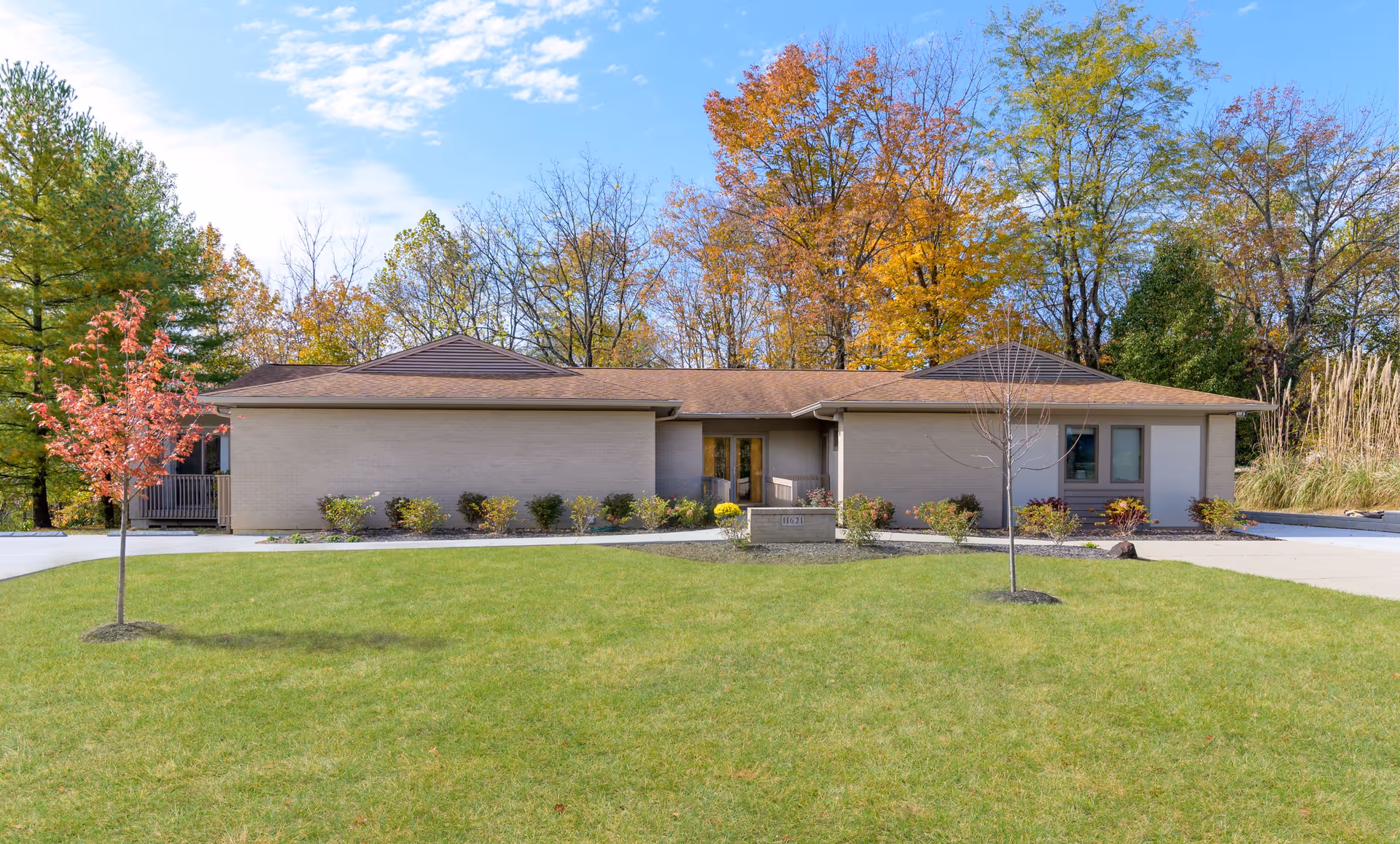 Single-story building with a brown roof and beige walls, surrounded by a well-maintained lawn and small trees with autumn foliage in the background under a partly cloudy sky.