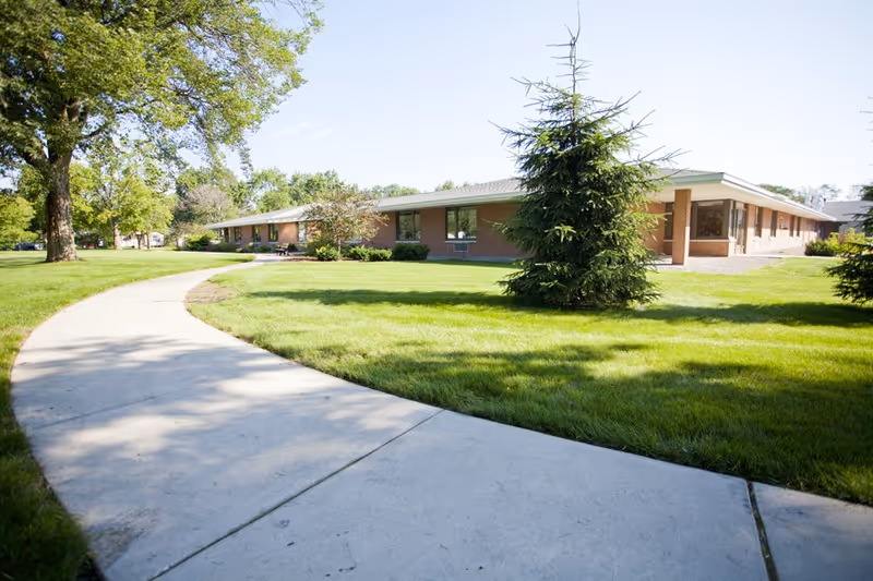 A single-story brick building with large windows surrounded by a well-maintained lawn and trees, with a curved concrete walkway leading towards the building under a clear blue sky.