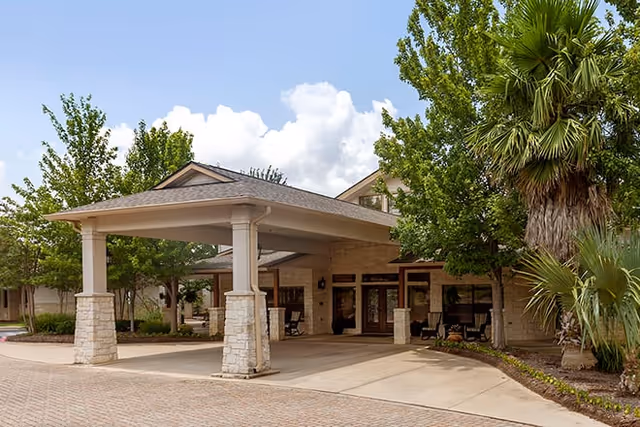 Covered porte-cochere entrance to a stone-faced building with outdoor seating and landscaped trees.