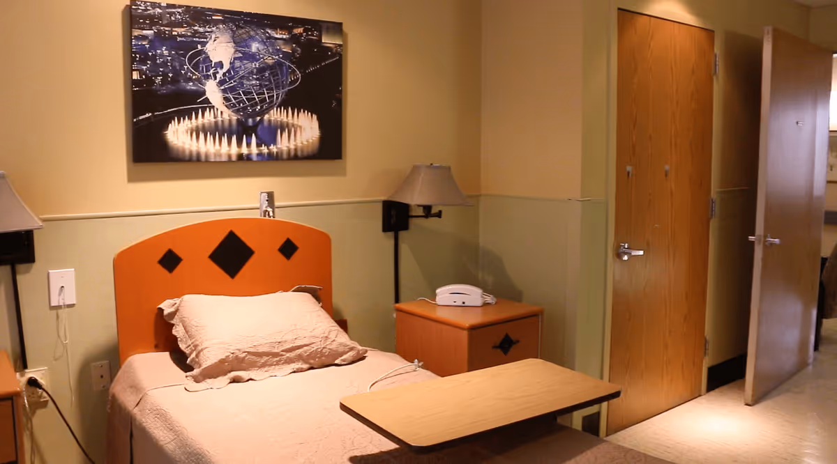 Single patient bedroom with a bed, headboard, bedside table and phone, overbed table, wall art, and an open door.