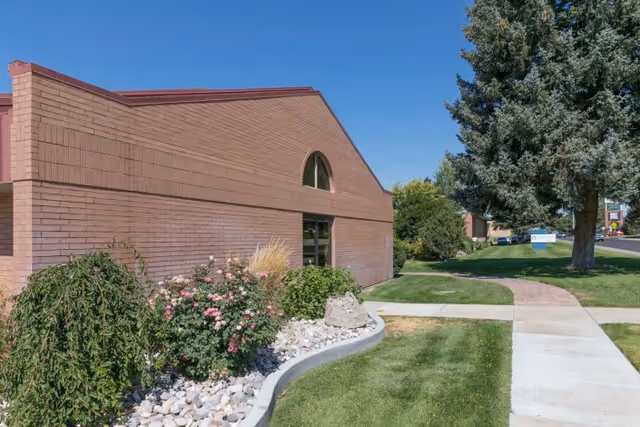 Brick exterior of a care center with landscaped flower beds, a sidewalk, and a large tree under a clear blue sky.