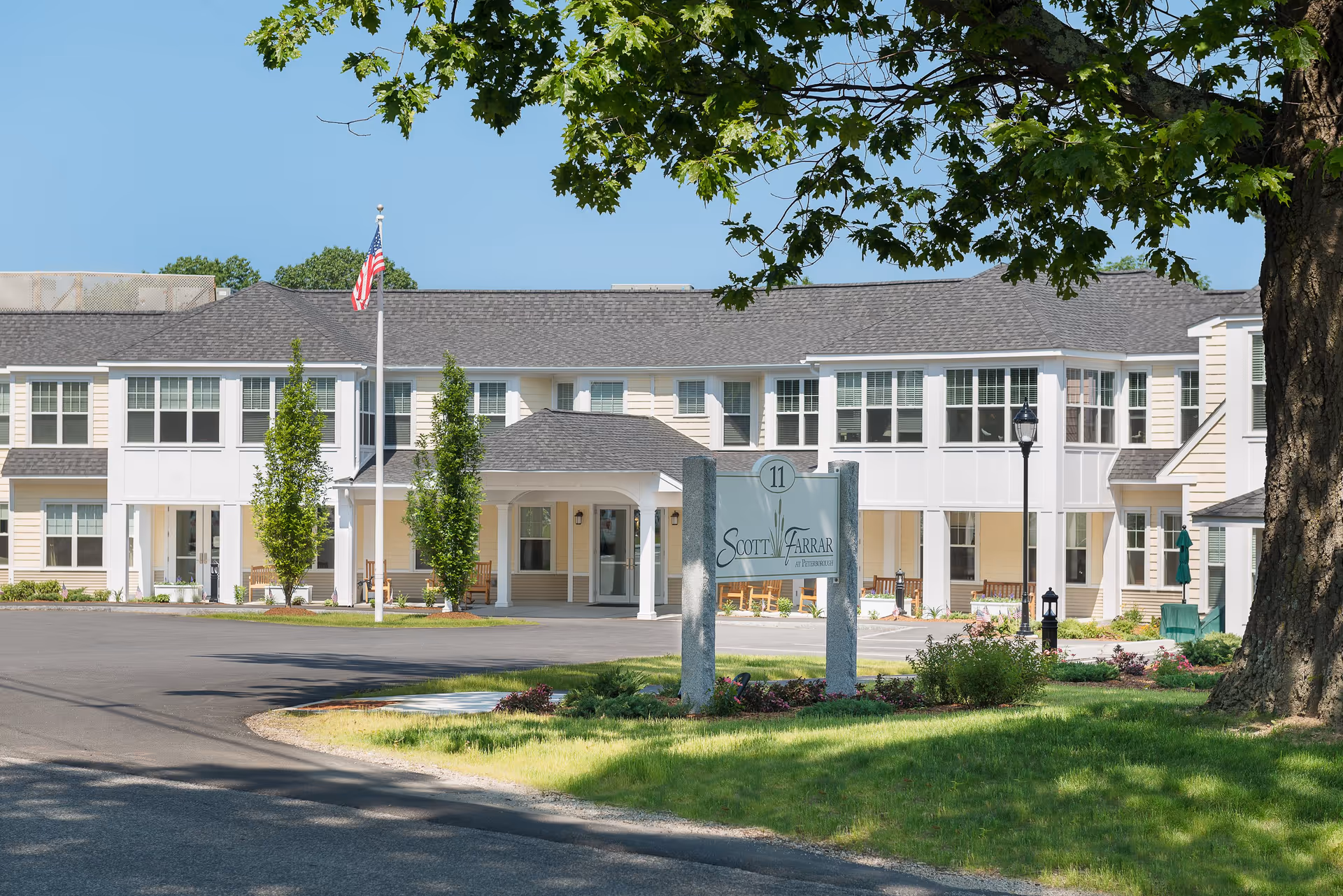 Exterior view of the Scott-Farrar at Peterborough senior living facility building with a driveway, green lawn, trees, and a sign displaying the facility name. The building is two stories with multiple windows and a covered entrance. An American flag is flying on a flagpole near the entrance.