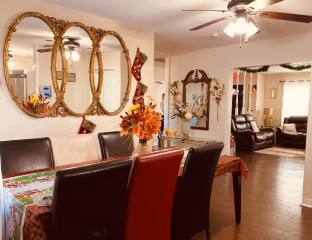 A dining room with a table covered by a festive tablecloth and a vase of autumn flowers in the center. There are six chairs around the table, with two red chairs and four black chairs. On the wall to the left, there are three ornate gold-framed oval mirrors. A ceiling fan with lights is above the table. In the background, there is a living room area with leather recliners and a window with curtains. The room is decorated with holiday stockings and garlands.