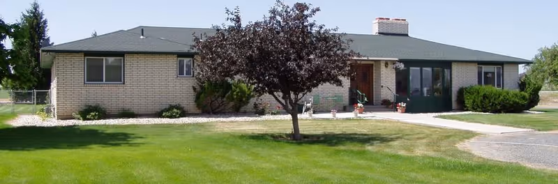 Single-story brick building with a dark roof, a central entrance with steps, a tree in front, and a well-maintained green lawn under a clear sky.