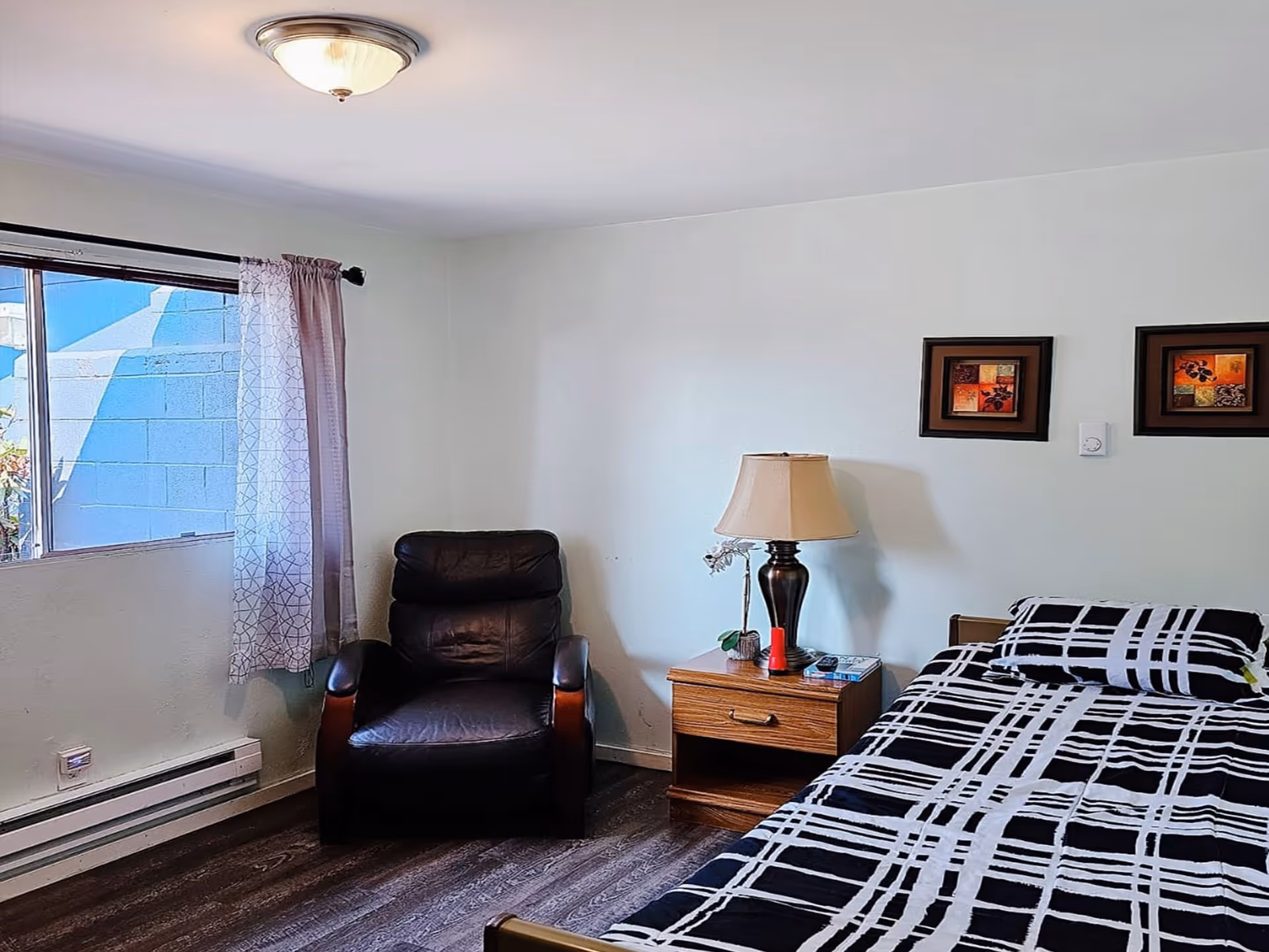 A tidy bedroom with a bed, nightstand, lamp, leather chair, and window curtains.