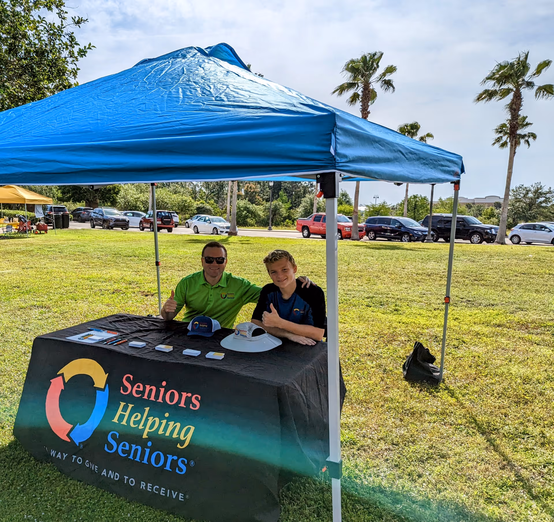 Two people sit at a table under a blue canopy outdoors; banner reads Seniors Helping Seniors.