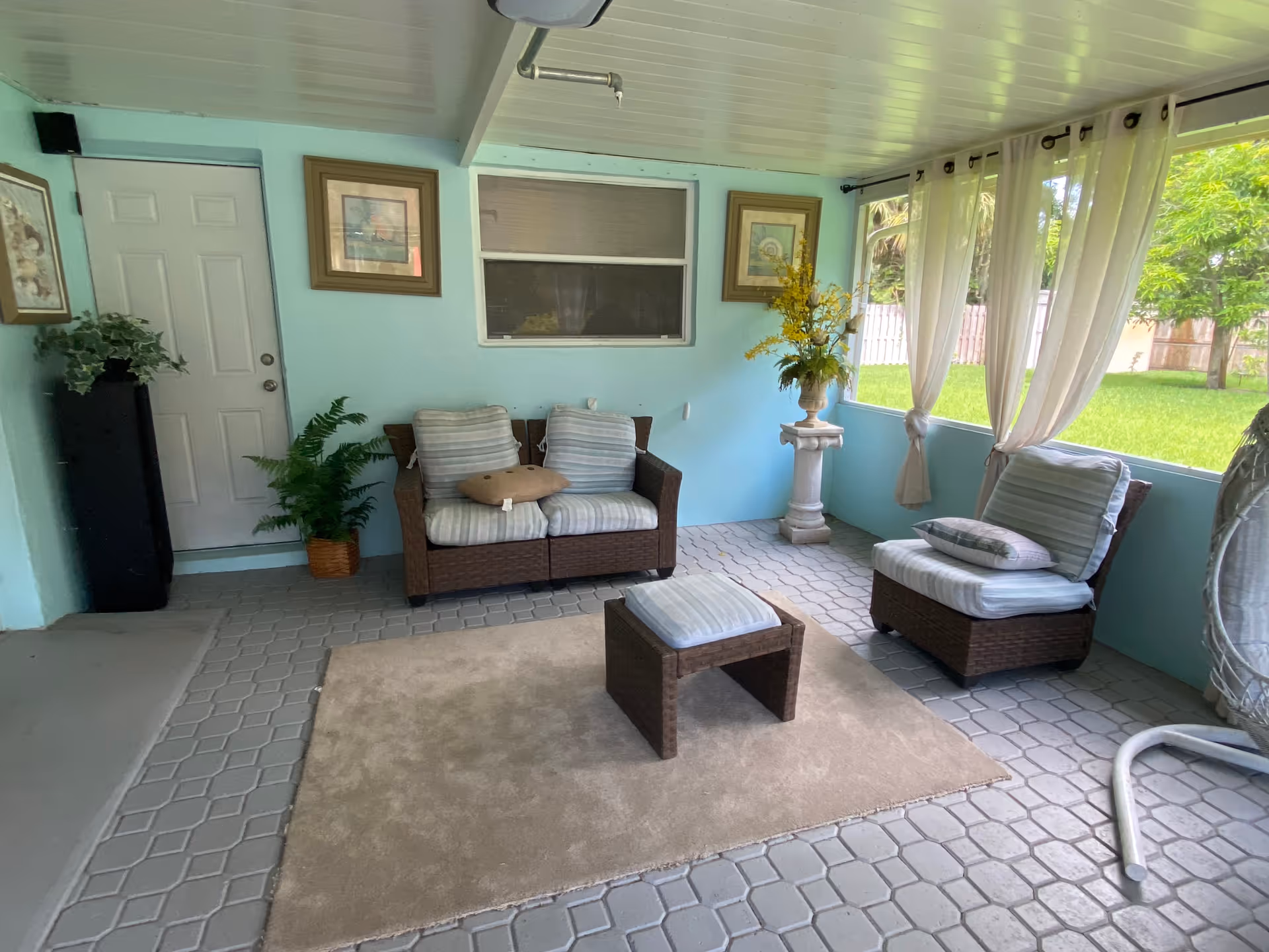 Covered patio with wicker seating, cushions, plants, and flowing curtains overlooking a green yard.