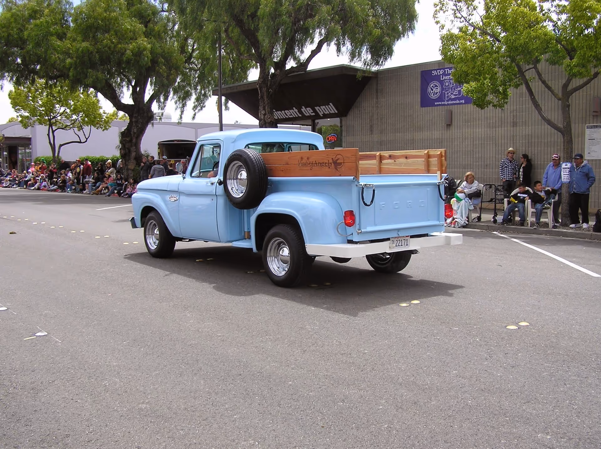 A light blue vintage pickup truck drives down a street with spectators along the curb.