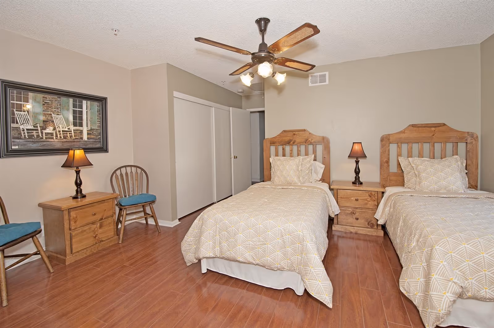 Twin bedroom with two wooden beds, nightstands and lamps, chairs, hardwood floor and a ceiling fan.