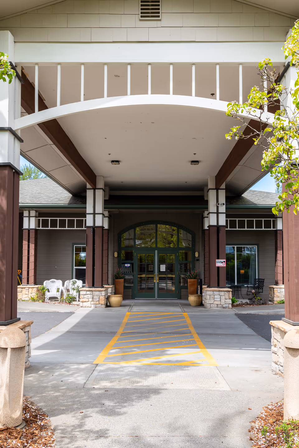 Covered entrance to Providence Brookside Manor with glass doors, yellow parking lines, and plants.