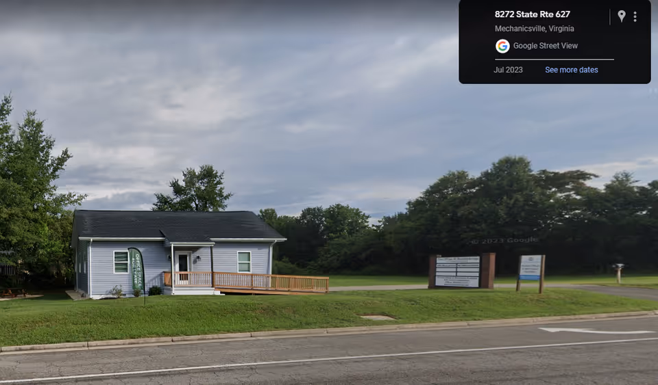 A small blue building with a wooden ramp and sign, framed by a grassy front yard.