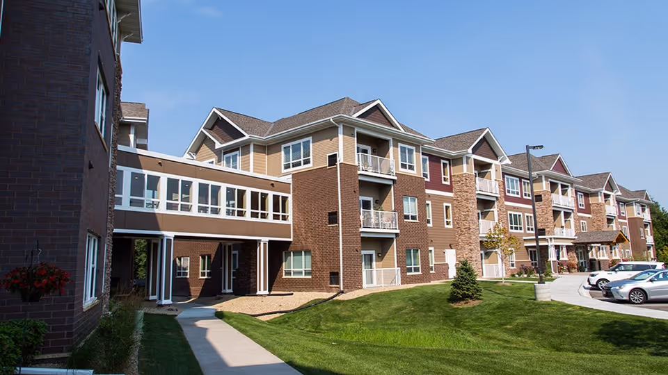 Exterior view of a multi-story senior living facility building with brick and beige siding, balconies, a covered walkway, green lawn, and parked cars under a clear blue sky.