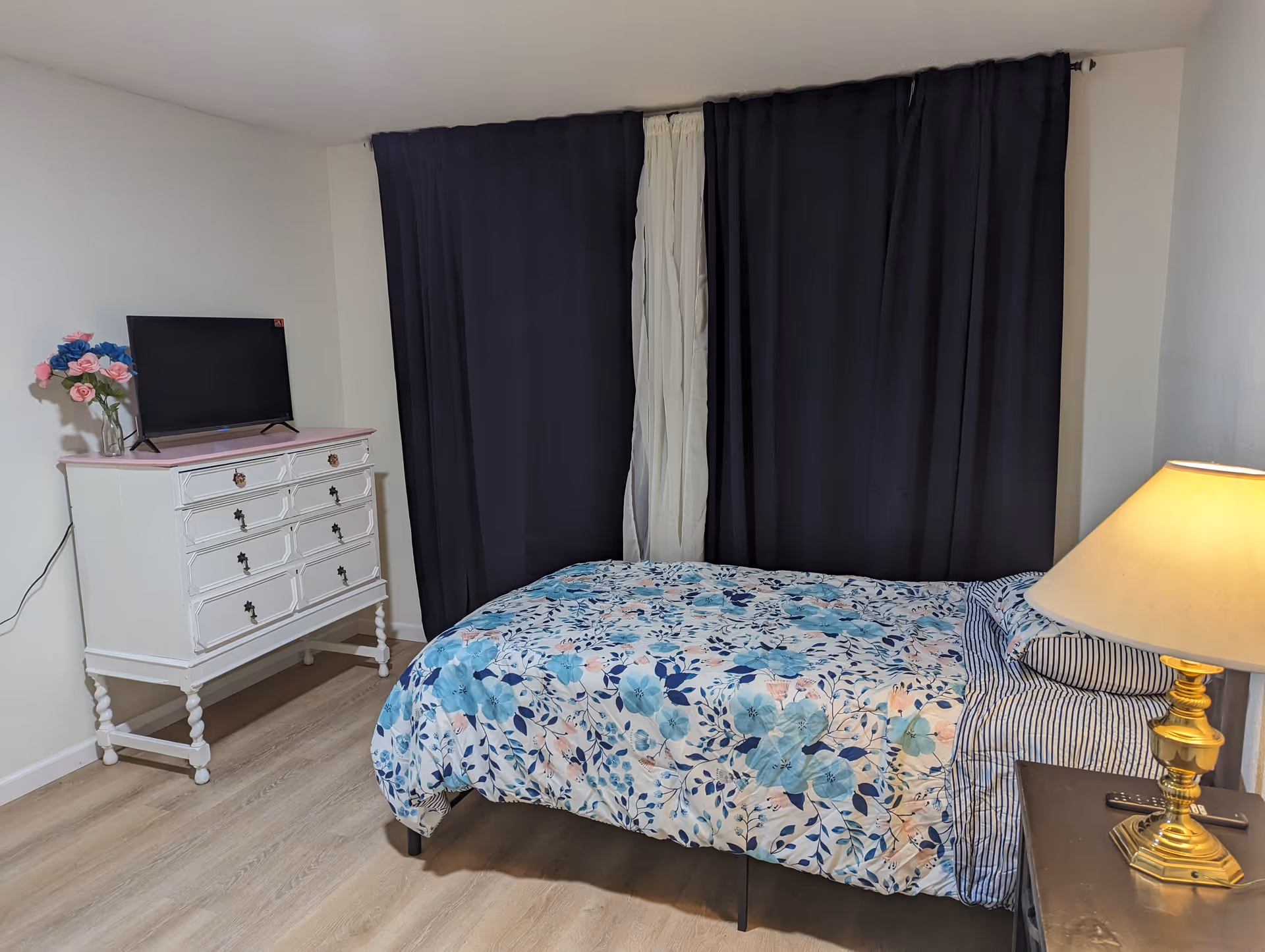 A bedroom with a floral bedspread, dresser with TV, and dark curtains.