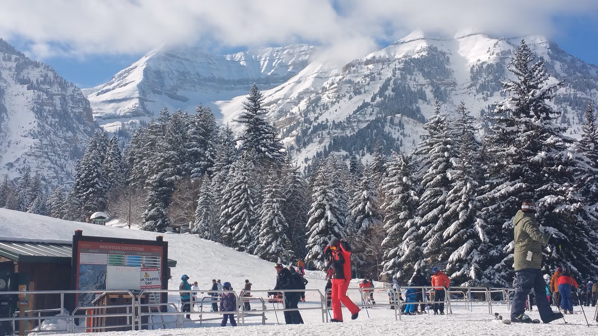Snow-covered mountains, evergreen trees, and people skiing on a sunny winter day.
