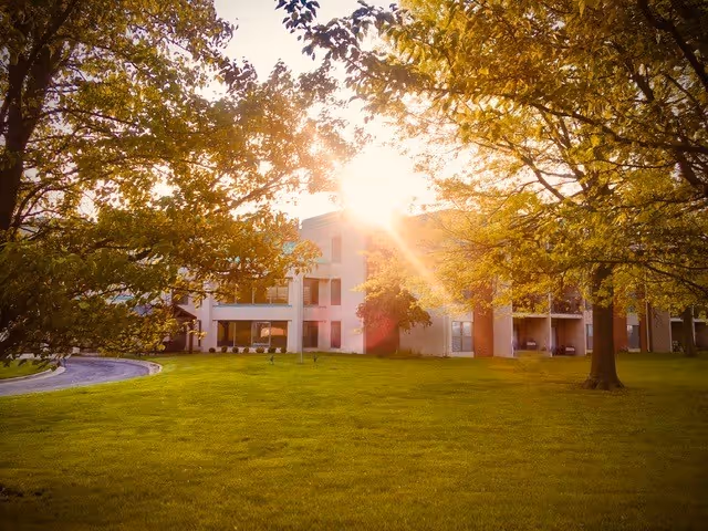 Exterior view of a multi-story senior living building with a sunlit lawn and trees in the foreground.