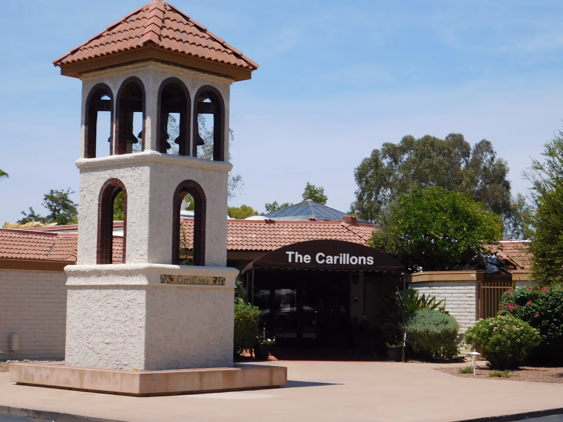 Entrance to The Carillons retirement community featuring a white stucco bell tower with arched openings and a red tile roof, surrounded by greenery and a clear blue sky.