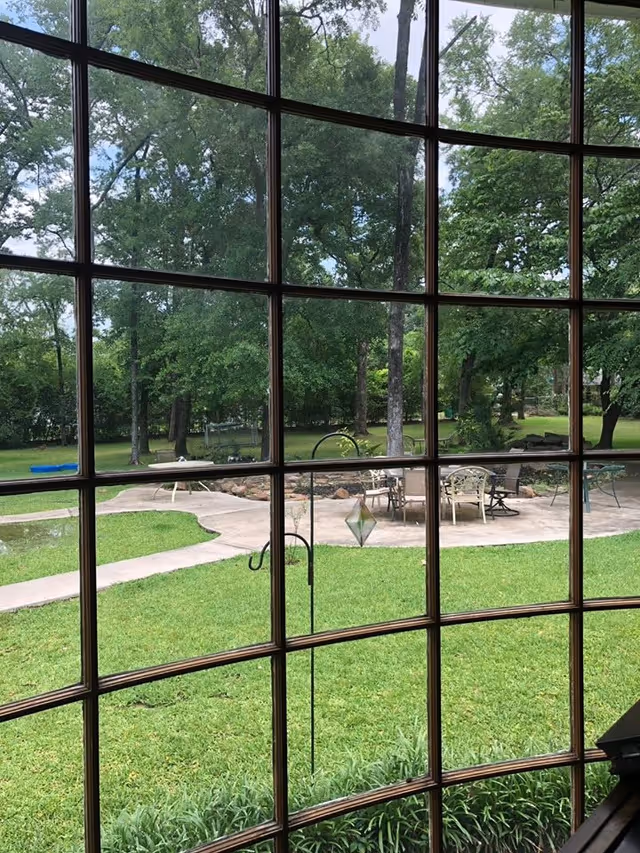 View through a multi-pane window onto a grassy lawn and patio with chairs surrounded by trees.