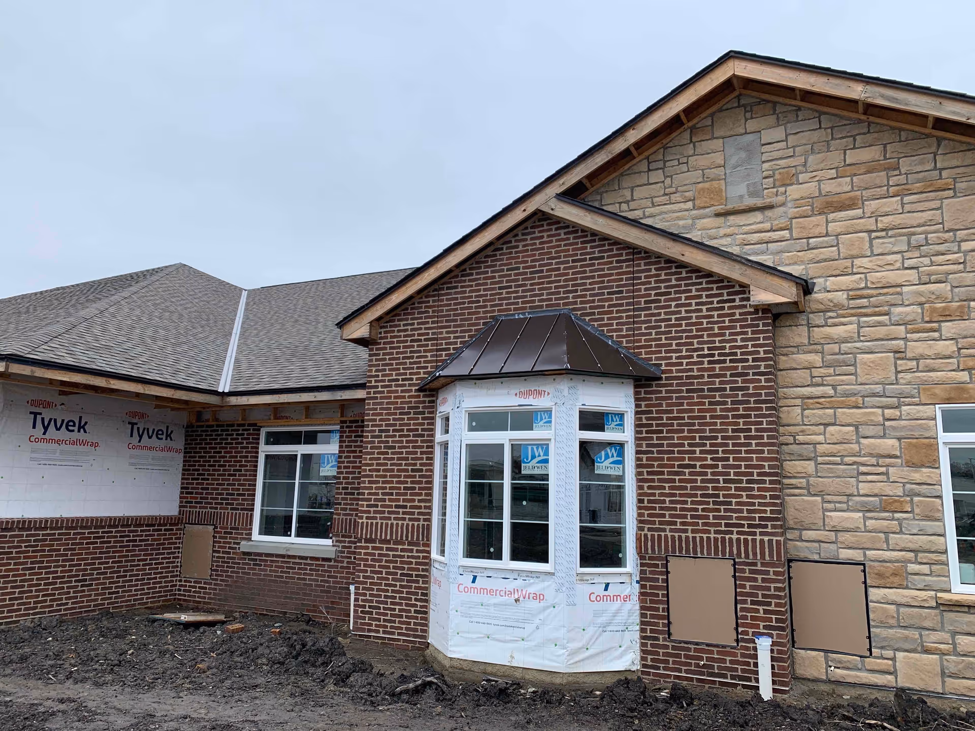 Exterior view of a building under construction with brick and stone walls, multiple windows, and a roof with shingles. The ground in front is muddy and unfinished.