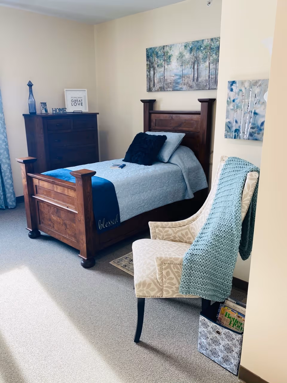 A cozy bedroom featuring a wooden bed with light blue bedding and a dark blue pillow. A wooden dresser with decorative items and framed quotes is against the wall. A patterned armchair with a light blue knitted throw draped over it sits near the bed. Two nature-themed paintings hang on the walls, and a small basket with books is placed beside the chair.
