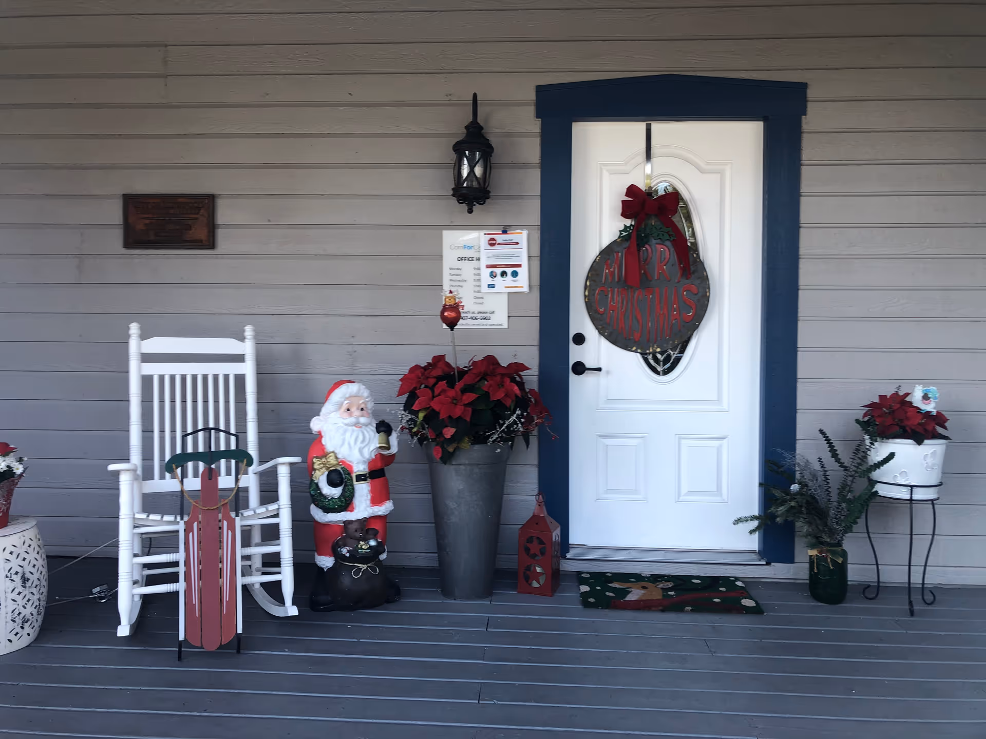 Front porch decorated for Christmas with Santa statue, rocking chair, poinsettias, and a wreath on door.