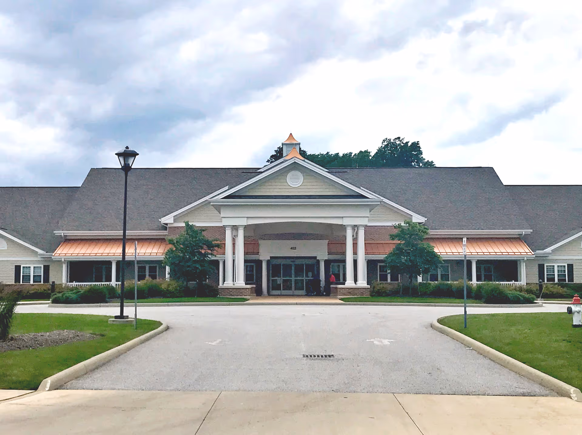 Front exterior view of Highland Pointe Health & Rehabilitation Center showing a large building with a peaked roof, white columns at the entrance, copper-colored awnings, a driveway, and landscaped greenery under a cloudy sky.